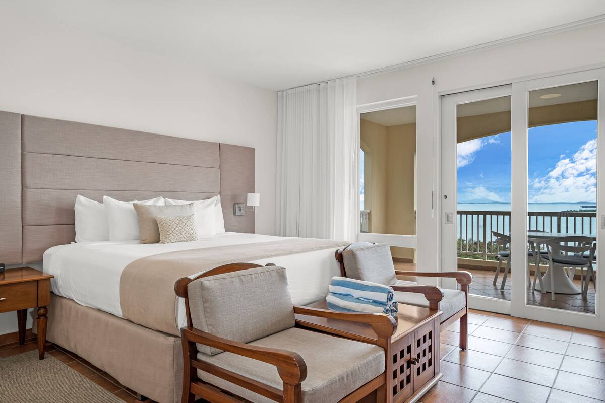 Habitación de hotel con una cama grande, una silla de madera, una mesita pequeña y un balcón con vistas.
