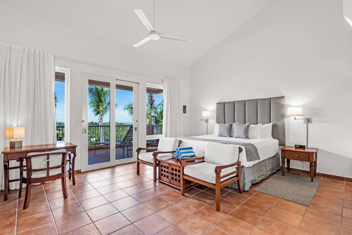 Un dormitorio con cama, escritorio, silla, mesita de noche y balcón con vista a palmeras.