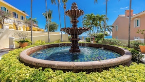 Una fuente circular con agua brotando del centro, rodeada por un jardín bien cuidado con diversas plantas y un cielo azul despejado al fondo.