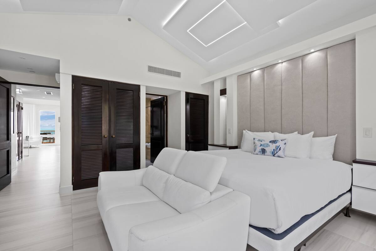 Un dormitorio moderno con una cama grande, un sofá blanco y vista al mar a través de la puerta abierta.