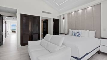 A modern bedroom with a large bed, a white sofa, and a view of the sea through the open door.
