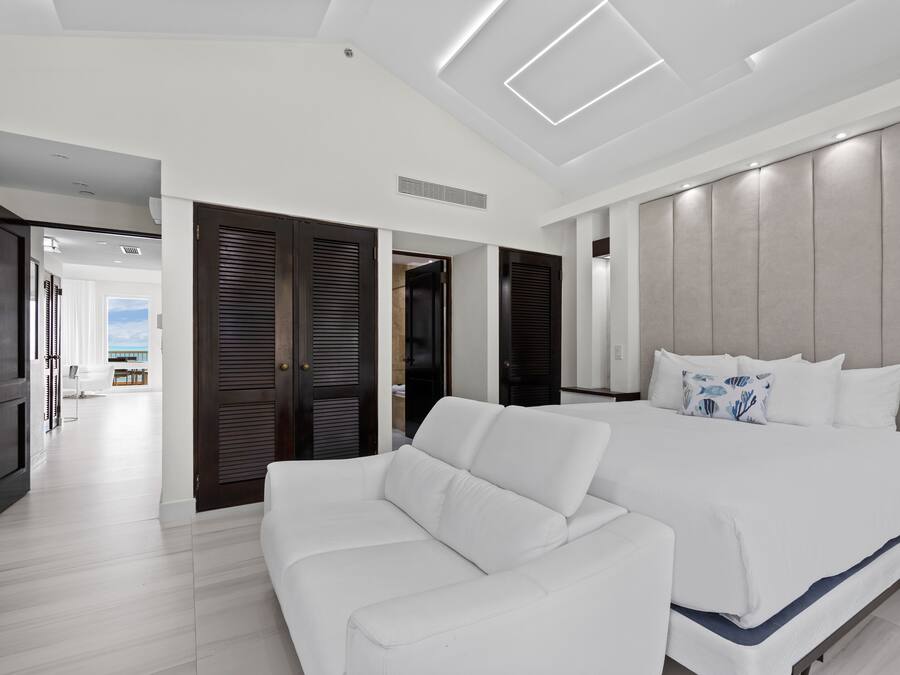 A modern bedroom with a large bed, a white sofa, and a view of the sea through the open door.