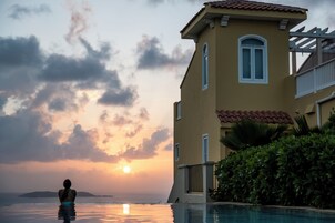 Une personne dans une piscine au coucher du soleil, avec un bâtiment en arrière-plan et une vue sur l’océan.