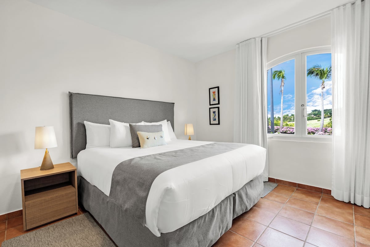 Habitación de hotel ordenada con una cama grande, una mesita de noche con lámpara y una ventana con cortinas.