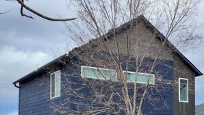 Una casa de dos pisos con fachada azul oscuro, una puerta de garaje visible y un pequeño árbol en la parte delantera.