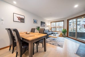 A modern living room with a dining area, large windows, and a view of the outside. - Heavenly Ski In/Out @ The Aspens w/ King Bed (Whistler)