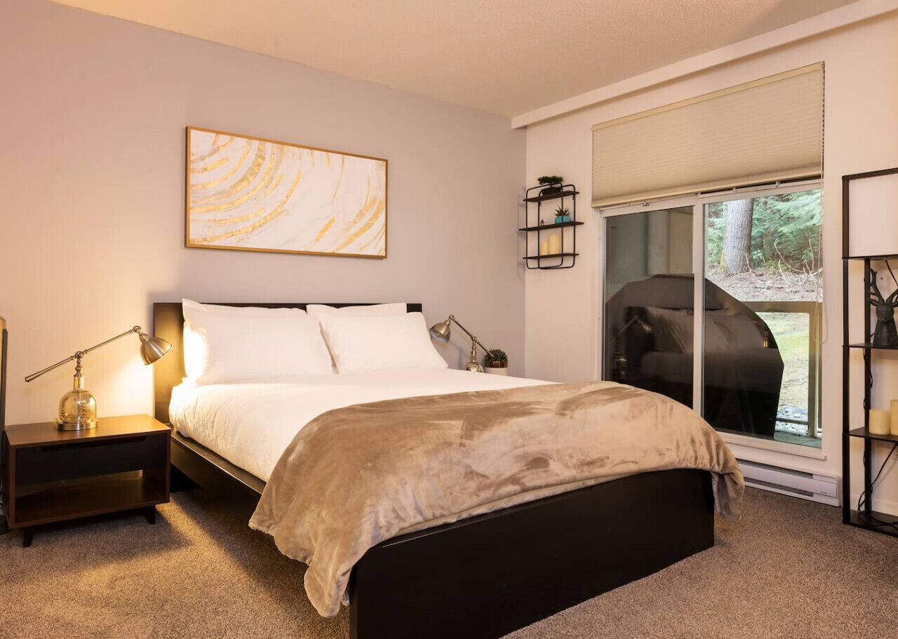 A bedroom with a bed, bedside table, lamp, and a window with a view of trees.