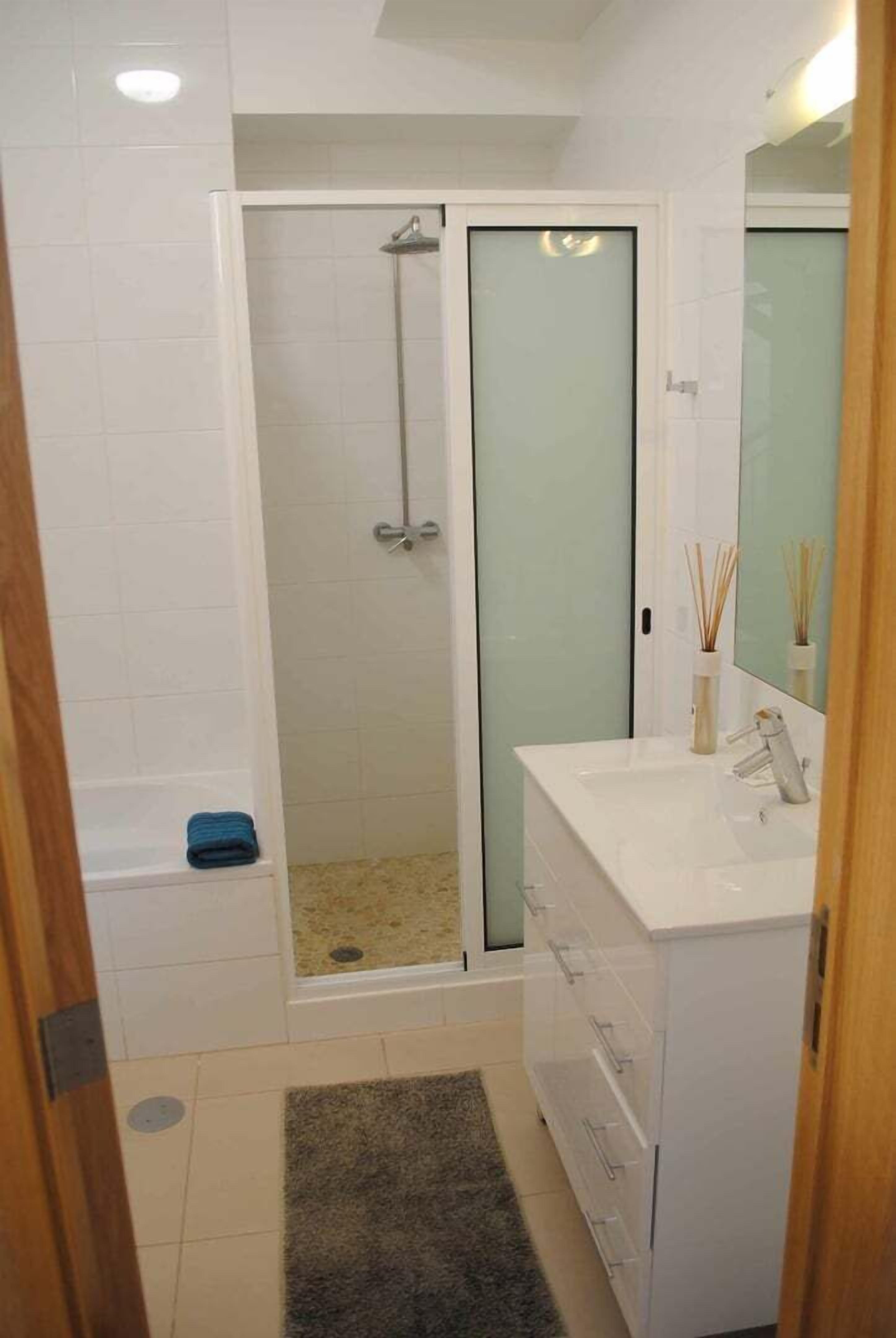 A bathroom with a glass-enclosed shower, a white sink with a mirror, and a small blue towel on the floor.