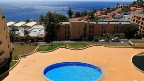 Piscina rodeada por um espaço pavimentado, com edifícios ao fundo.