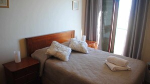 Cama bem arrumada com cabeceira de madeira, duas almofadas fofas e uma toalha dobrada sobre um edredom bege. Tem uma mesinha de cabeceira de madeira com uma vela branca e uma moldura com foto na parede ao lado da cama.