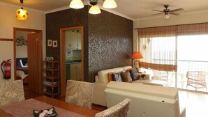 A living room with a sofa, a dining table with a placemat, a ceiling fan, and a window with blinds. - Magos Sunrise - Apt with sea view (Santa Cruz)