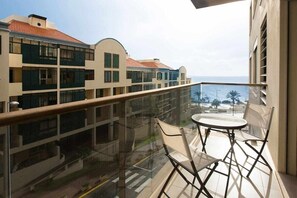 Un balcone con ringhiera in vetro, un tavolino rotondo e due sedie. Sullo sfondo, edifici a più piani con balconi.