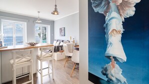 A modern kitchen with a large window, a dining area with white chairs, and a painting of an angel on the wall. - Barreirinha II Near Old Town With Nice Ocean Views (Funchal)