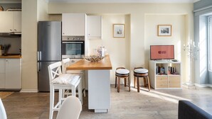 A modern kitchen with a refrigerator, oven, and dining area with stools. There is a TV on a stand with a 'GUEST READY' sign. - Barreirinha II Near Old Town With Nice Ocean Views (Funchal)