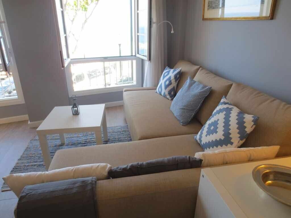 A modern living room with a beige sofa, patterned cushions, a small white table, and a view through a large window.
