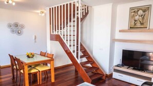 A modern living room with a wooden staircase, a dining table with chairs, a flat-screen TV, and a painting on the wall. - Rosalina's Apartment (Caniço)