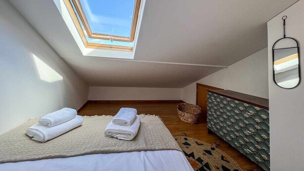 A small, modern bedroom with a skylight, a bed with white linens, a wooden headboard, and a wall-mounted mirror. - Rosalina's Apartment (Caniço)