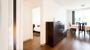 Une chambre d’hôtel moderne avec un plancher en bois, une armoire en bois foncé, un lit avec une literie blanche et un coin repas comprenant une table et des chaises.