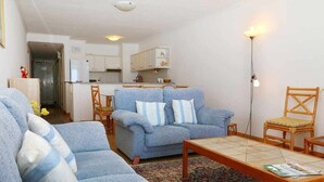 Ein Wohnzimmer mit einer Couch, einem Couchtisch und einem Sessel. Im Hintergrund ist eine Küche zu sehen.