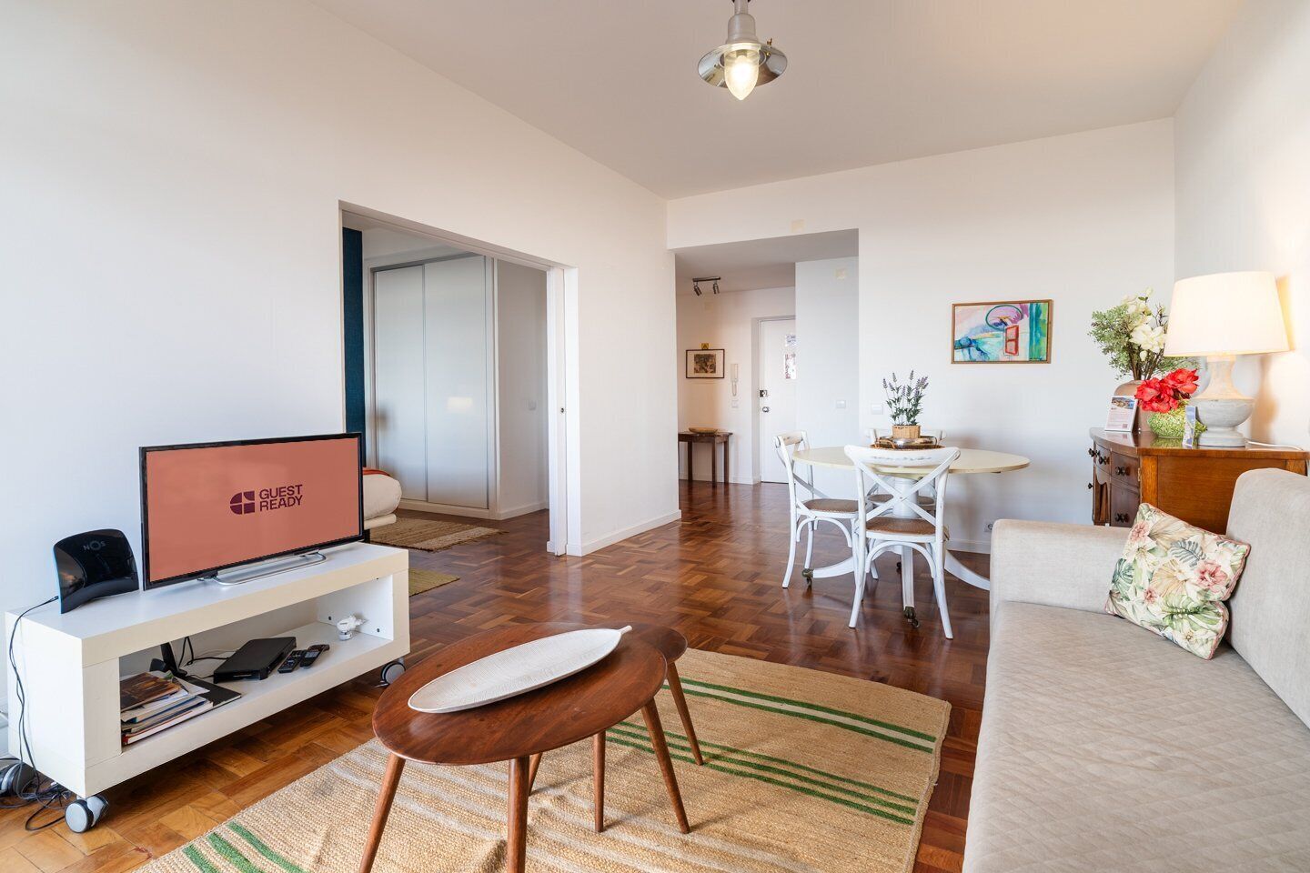 A modern living room with a flat-screen TV, a sofa, a dining table with chairs, and a kitchen area in the background.
