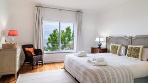 A bedroom with a large bed, a chair, a nightstand, a lamp, and a window with a view of trees. - Vila da Falésia-With Pool and views (Caniço)