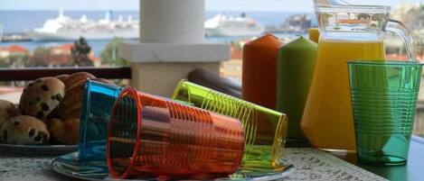 Una mesa con vasos de colores, una jarra de jugo de naranja y un plato de galletas en un balcón con vistas a un puerto y barcos.