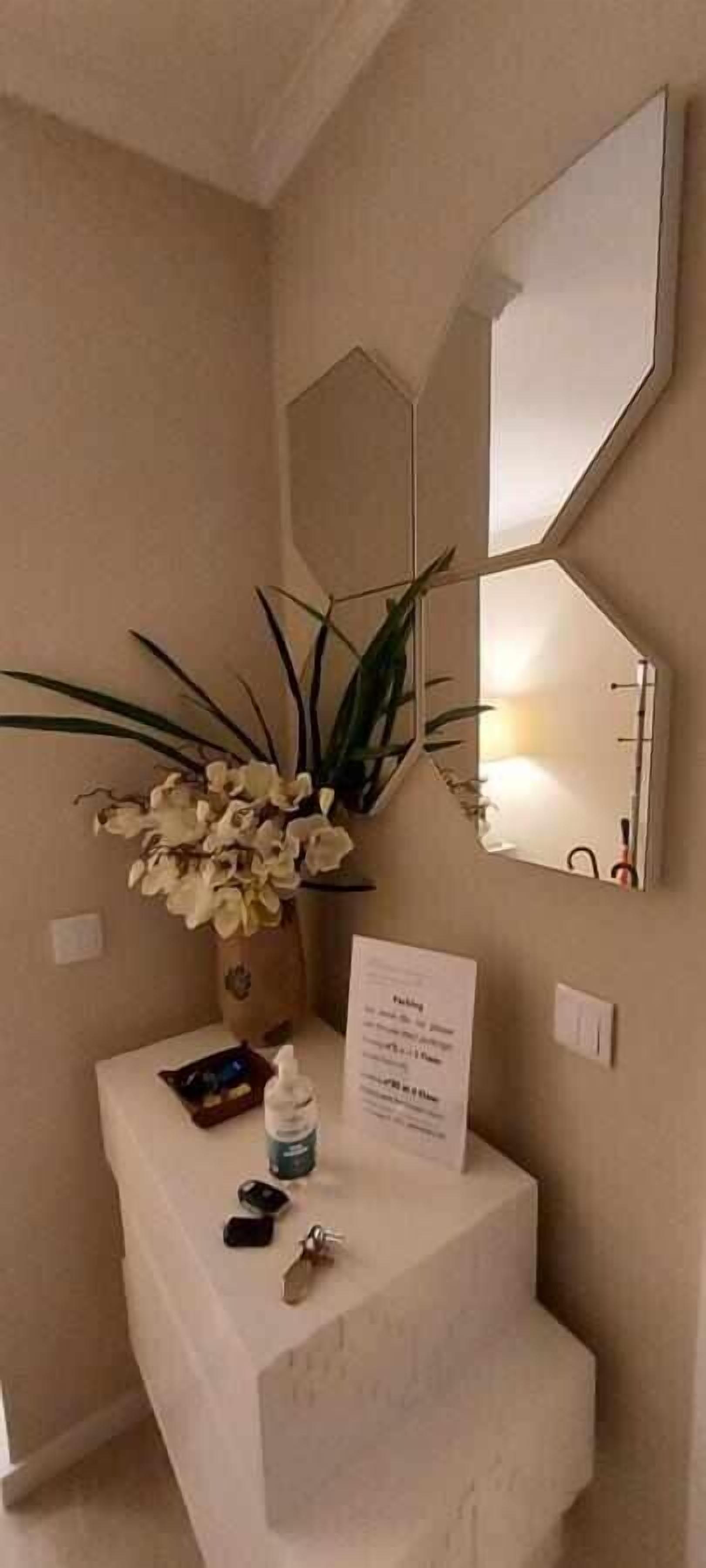 A bathroom with a white vanity, a mirror, a vase of flowers, a bottle of hand sanitizer, and a sign with text.