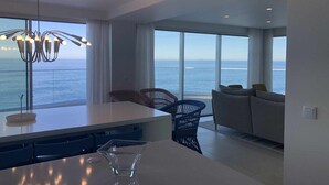 Ein modernes Wohnzimmer mit großem Fenster mit Meerblick, einer Essecke mit weißem Tisch und Stühlen sowie einer stilvollen Pendelleuchte.
