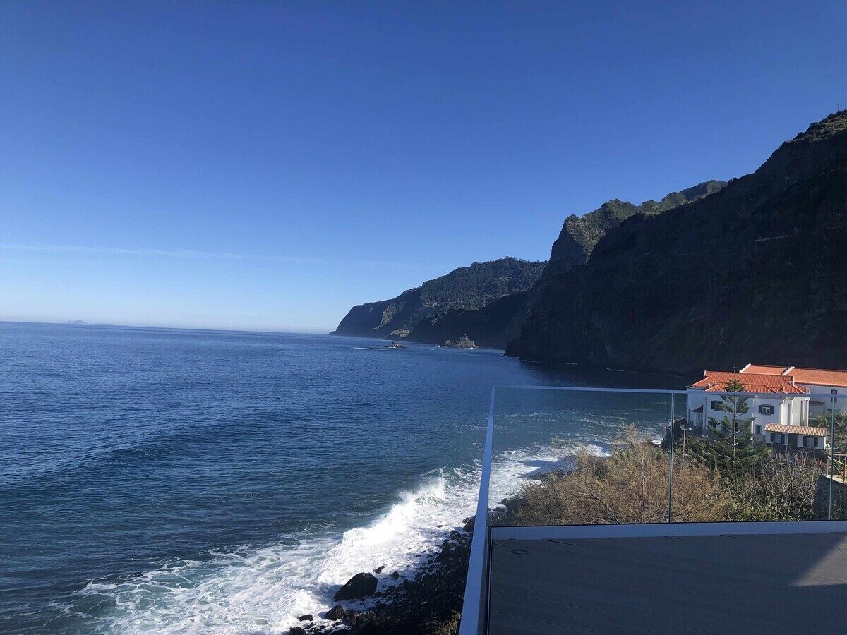 A coastal view with a building adjacent to the sea, featuring a balcony with a clear view of the ocean and cliffs.