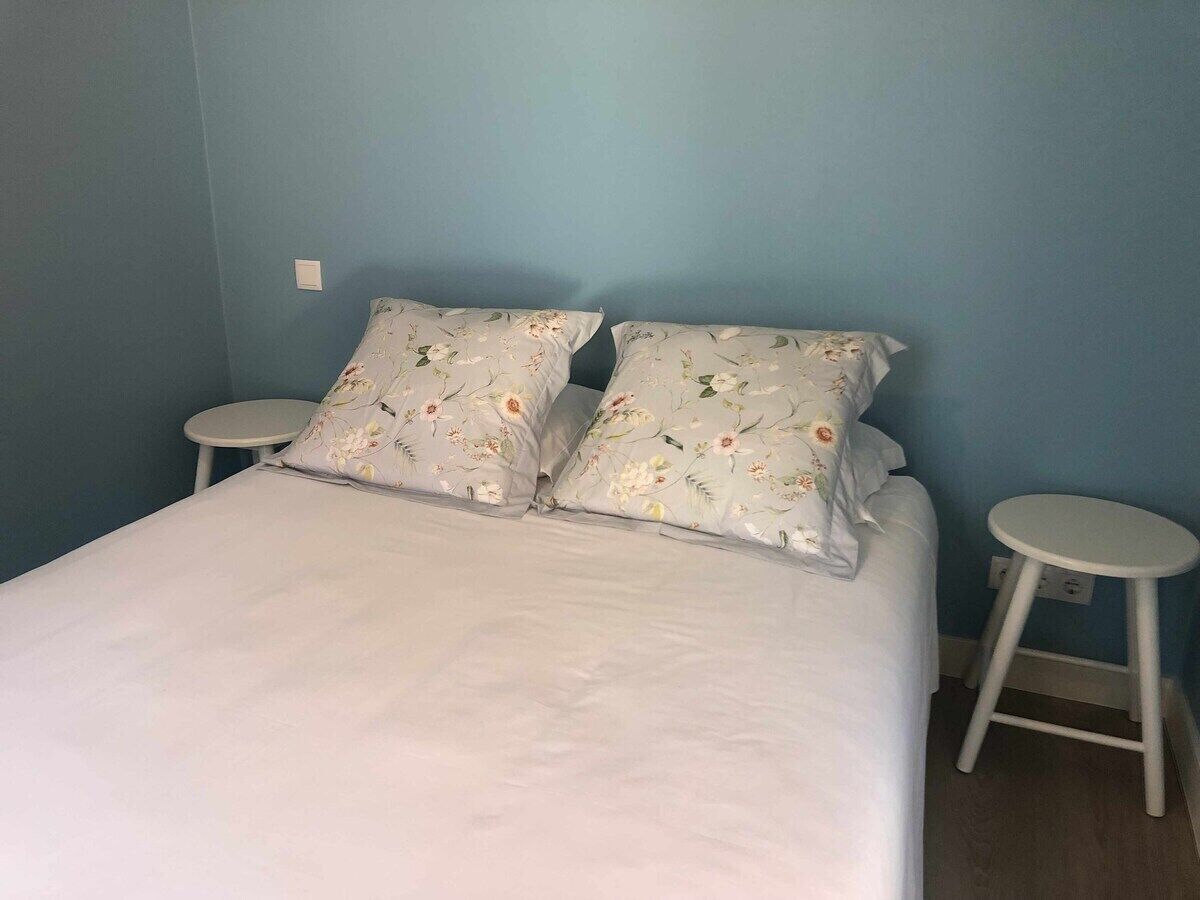 A bed with floral-patterned pillows, two white round side tables, and a blue wall.