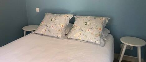 A bed with floral-patterned pillows, two white round side tables, and a blue wall.
