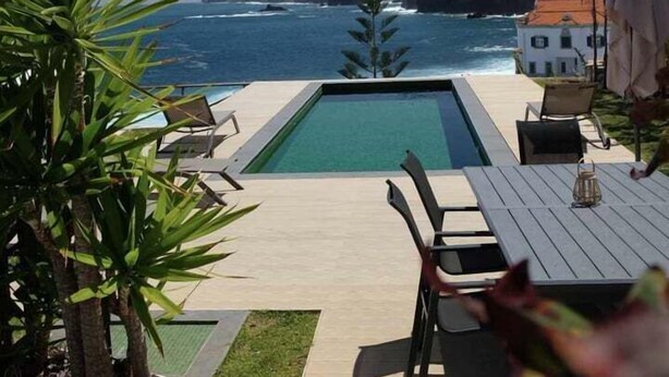 Ein Poolbereich mit Blick auf den Ozean, ein weißes Gebäude und eine Terrasse mit Stühlen und Tisch.