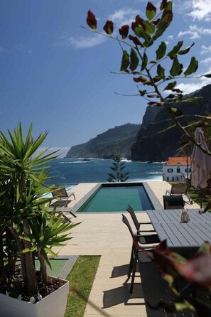 Ein Poolbereich mit Blick auf den Ozean, ein weißes Gebäude und eine Terrasse mit Stühlen und Tisch.