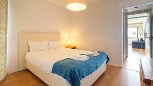 Ein ordentlich bezogenes Bett mit weißen Leinentüchern und einer blauen Decke, eine Nachttischlampe und ein Blick in einen anderen Raum mit einem Sofa.