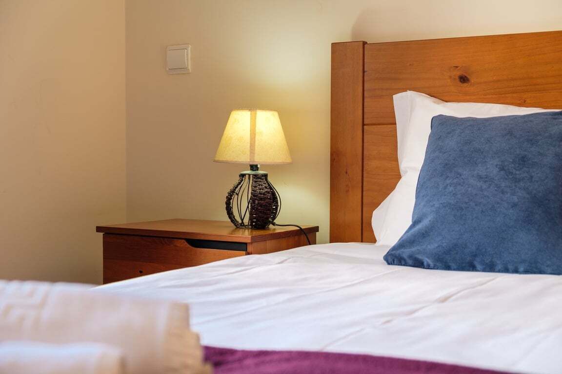 A neatly made bed with white sheets and a blue pillow, a wooden nightstand with a lamp, and a wooden headboard.