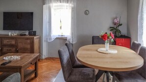 A living room with a wooden dining table, grey upholstered chairs, a flat-screen TV on a wooden cabinet, a window with sheer curtains, and a vase with flowers on the table.