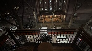 Un balcón con sillas y mesas, con vistas a una calle donde se ven coches aparcados y un edificio con ventanas iluminadas.