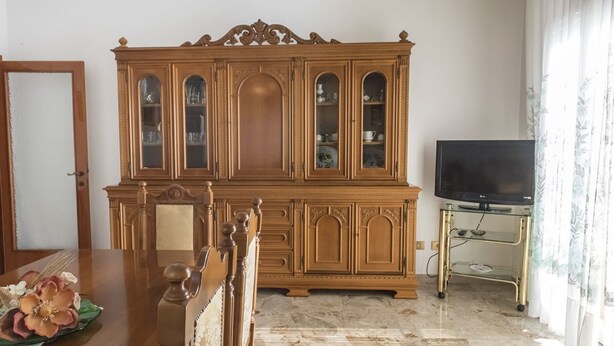 Un comedor con una mesa de madera, sillas, un mueble bar con puertas de vidrio y un televisor sobre un soporte.