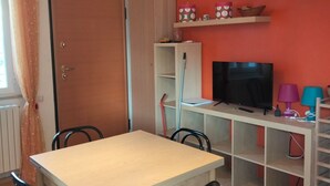 Una habitación con una mesa de madera, sillas, un televisor, estantes con objetos y una puerta.