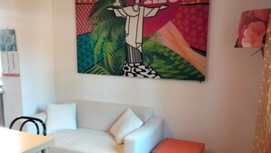 Una habitación con un sofá blanco, una pequeña mesa y una silla. Un cuadro colorido adorna la pared sobre el sofá.