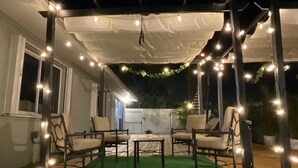An outdoor patio with a wooden deck, a covered seating area with chairs and a table, and string lights. - Dream Vacation House 4B/2B Miami, FL (Hialeah)
