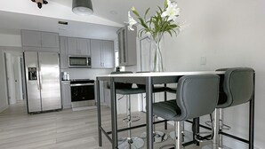 A modern kitchen with stainless steel appliances, a white dining table with bar stools, and a vase with flowers on the table. - Dream Vacation House 4B/2B Miami, FL (Hialeah)