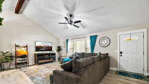 A living room with a ceiling fan, a sofa, a TV, a clock, and a door. - The Happy Place: 3BR in Quiet Neighborhood (Douglasville)