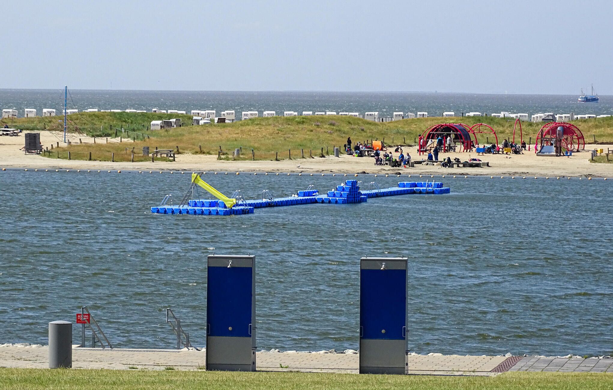 Ett område vid vattnet med en brygga som sträcker sig ut i vattnet, en strand med folk och en röd byggnad, samt ett fartyg i fjärran.