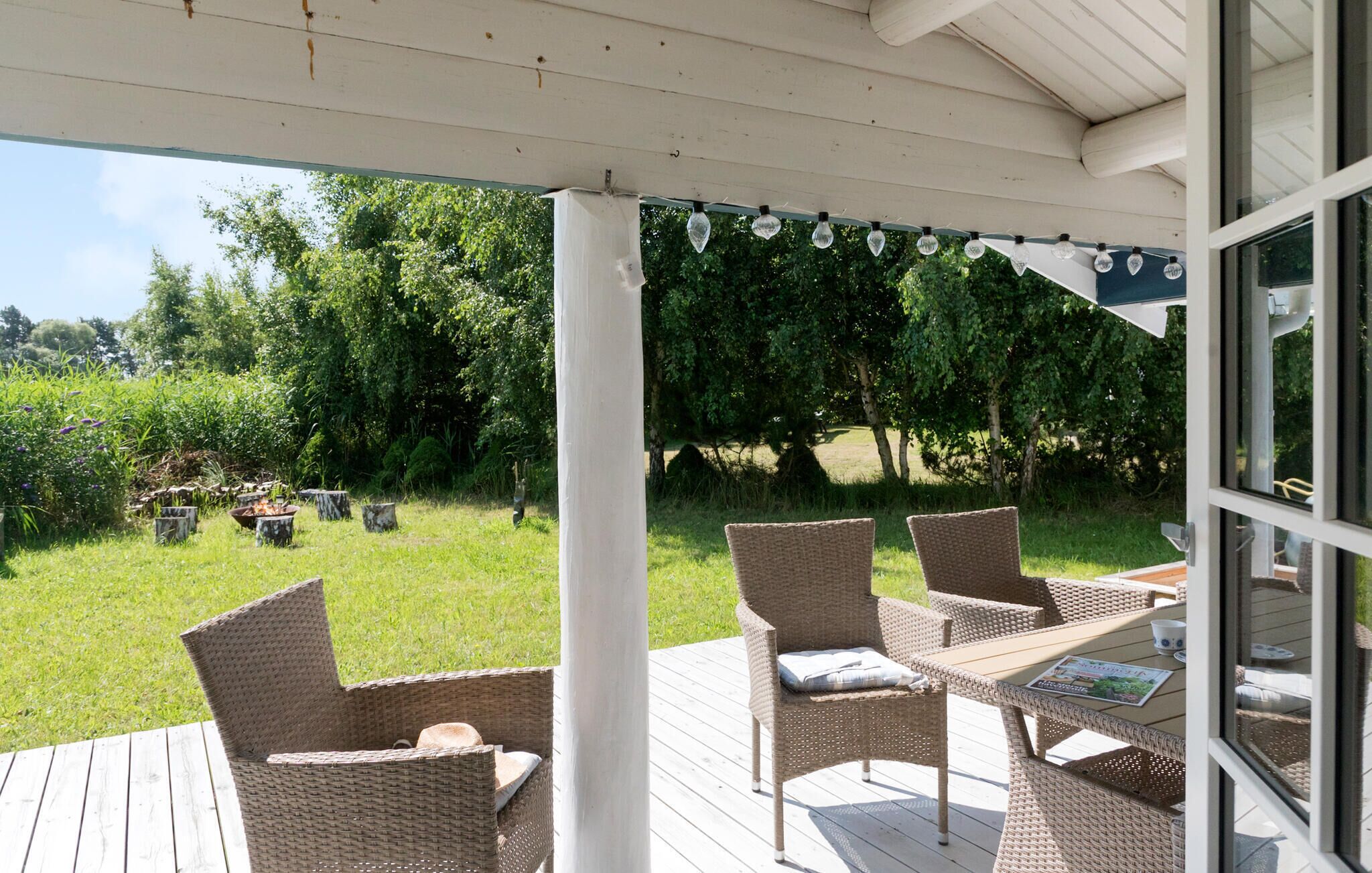 An outdoor patio with wicker furniture and a table, overlooking a grassy area with trees and a garden.