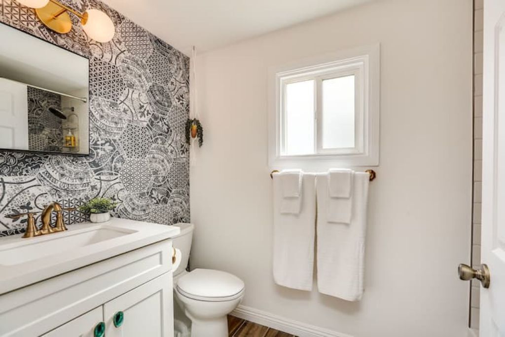 Une salle de bain avec un lavabo blanc, un robinet doré et un mur à motifs. Il y a une fenêtre et une serviette est accrochée à la porte.