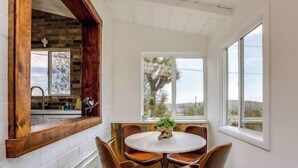 A dining area with a round table and six chairs, a window with a view of trees, and a brick wall with a wooden beam. - J.T. Cottage (Yucca Valley)