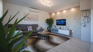Una sala de estar moderna con un sofá gris, una mesa de centro redonda, un televisor de pantalla plana grande montado en una pared texturizada y una planta en maceta.