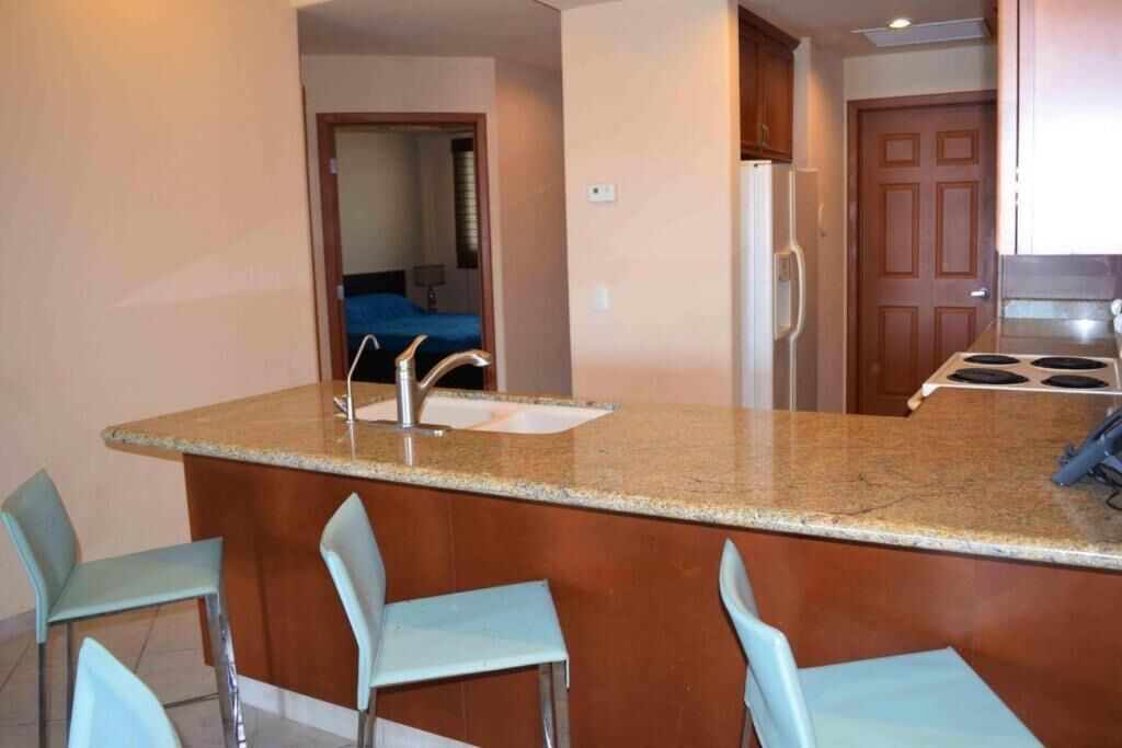 A kitchen with a granite countertop, a sink, and a refrigerator. There are four light blue chairs around the counter.