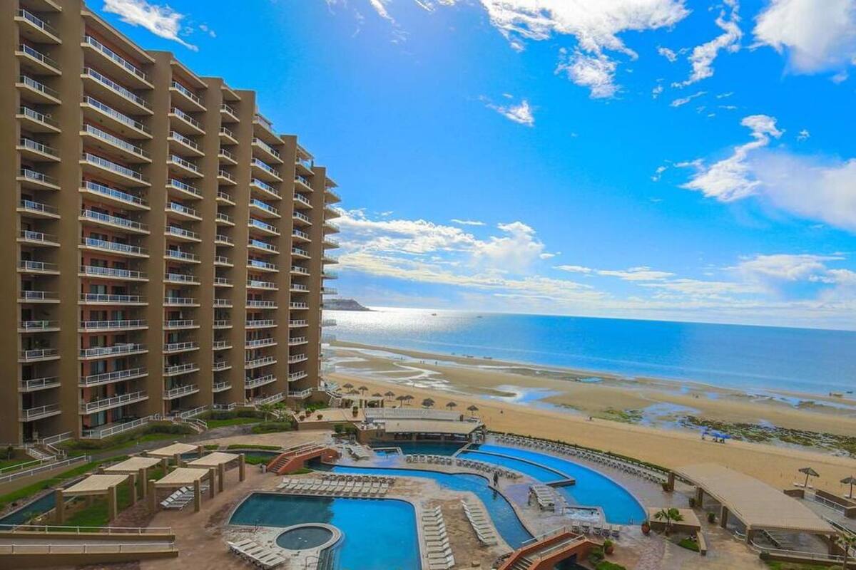 A high-rise hotel with multiple balconies overlooking a beach and a pool area with multiple pools and sun loungers.